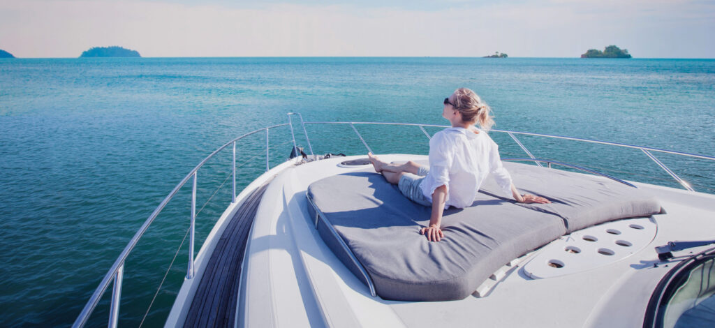 A person relaxes on the sun deck of a yacht, contemplating travel expenses while sitting on a cushion and gazing at the vast, calm ocean. The sky is clear, and small islands are visible in the distance.