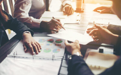 People are meeting around a table with papers and charts, electing the best strategies for residential rental property investments. Hands are pointing and writing notes. Warm lighting suggests a collaborative atmosphere, focusing on analytical work and discussion.