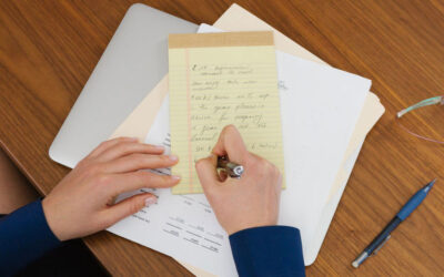 A person is writing on a yellow notepad with a pen, on a table with scattered papers and a closed laptop. Nearby are a pair of glasses and a blue pen. The scene suggests a workspace or study area.