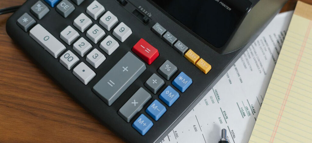 A calculator with various colored buttons is placed on top of several papers, including a financial statement and a yellow notepad. A black pen is also visible at the bottom of the image on the wooden surface.