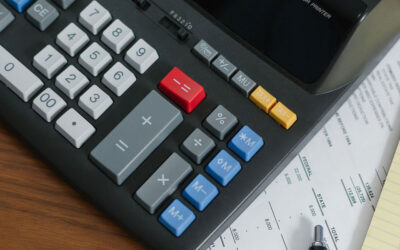 A calculator with various colored buttons is placed on top of several papers, including a financial statement and a yellow notepad. A black pen is also visible at the bottom of the image on the wooden surface.