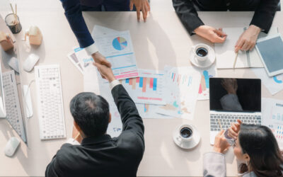 Aerial view of a business meeting with four people around a table, surrounded by laptops, coffee cups, and papers filled with graphs and charts. Two are shaking hands over a buy-sell agreement, signaling a promising collaboration.