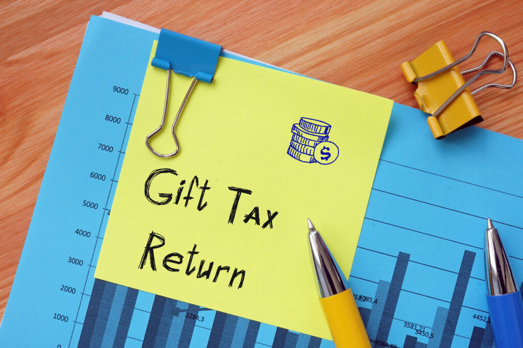 A blue chart with vertical bars is on a wooden surface. A yellow sticky note reads "File Gift Tax," featuring a dollar sign and coin illustration above. Two pens and colorful binder clips are scattered on the paper.