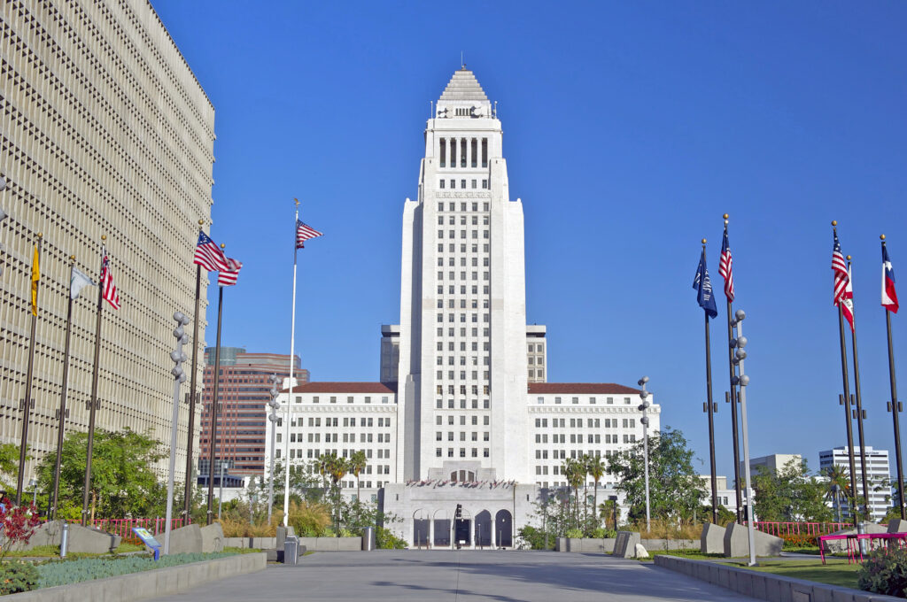 The image reveals a large white building with a tall central tower, flanked by several American flags. The clear blue sky enhances the urban setting, with other structures in the backdrop. This emblematic scene subtly underscores the economic pulse of California’s Pass-Through Entity tax landscape.