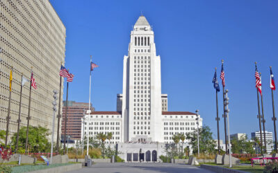The image reveals a large white building with a tall central tower, flanked by several American flags. The clear blue sky enhances the urban setting, with other structures in the backdrop. This emblematic scene subtly underscores the economic pulse of California’s Pass-Through Entity tax landscape.