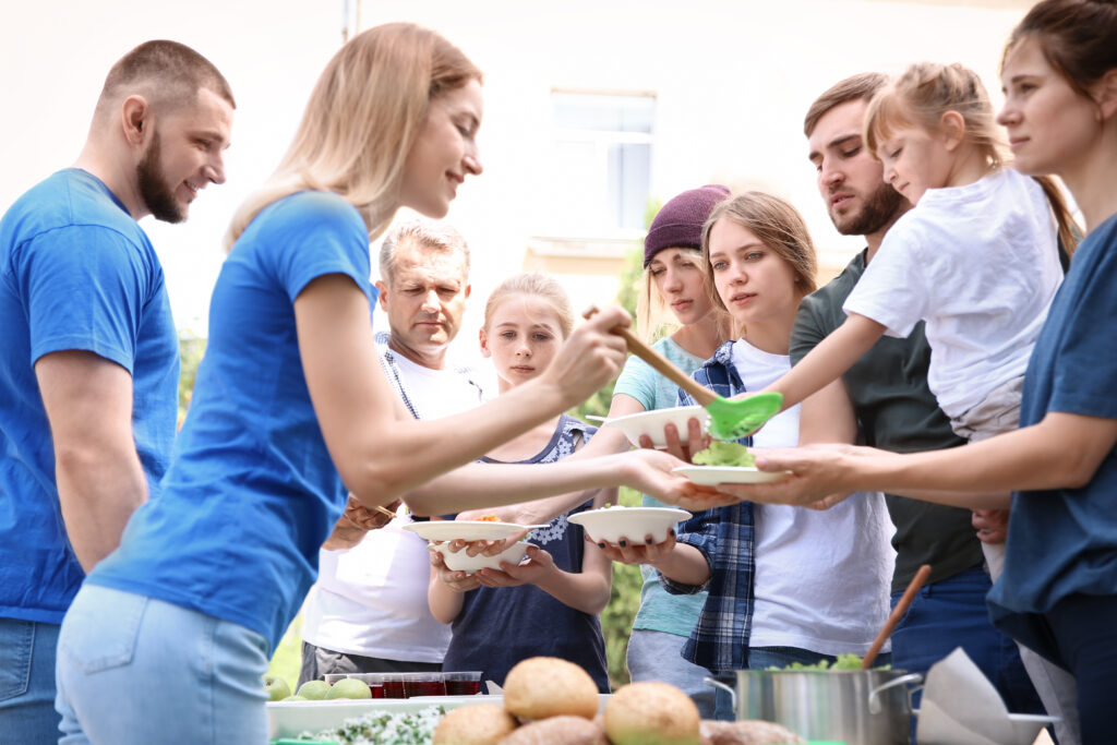 At a casual outdoor gathering, people serve and receive food from a table laden with salads. A woman in a blue shirt cheerfully serves while engaging with children and adults alike, embodying the spirit of charitable giving in this warm, social setting.