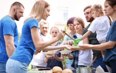 At a casual outdoor gathering, people serve and receive food from a table laden with salads. A woman in a blue shirt cheerfully serves while engaging with children and adults alike, embodying the spirit of charitable giving in this warm, social setting.