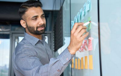 In an office setting, a man in a gray shirt focuses intently as he writes on a glass board covered with colorful sticky notes. He's clearly organizing ideas or tasks for a client memo, ensuring everything aligns perfectly for the small business’s big concerns.