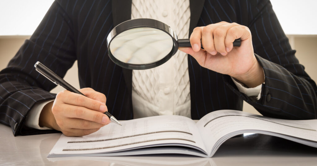A person in a pinstripe suit uses a magnifying glass to closely examine an open book or document, perhaps preparing for an IRS partnership audit. They hold a pen, poised to take notes or make annotations on the pages. The scene suggests meticulous study of tax rules and regulations.
