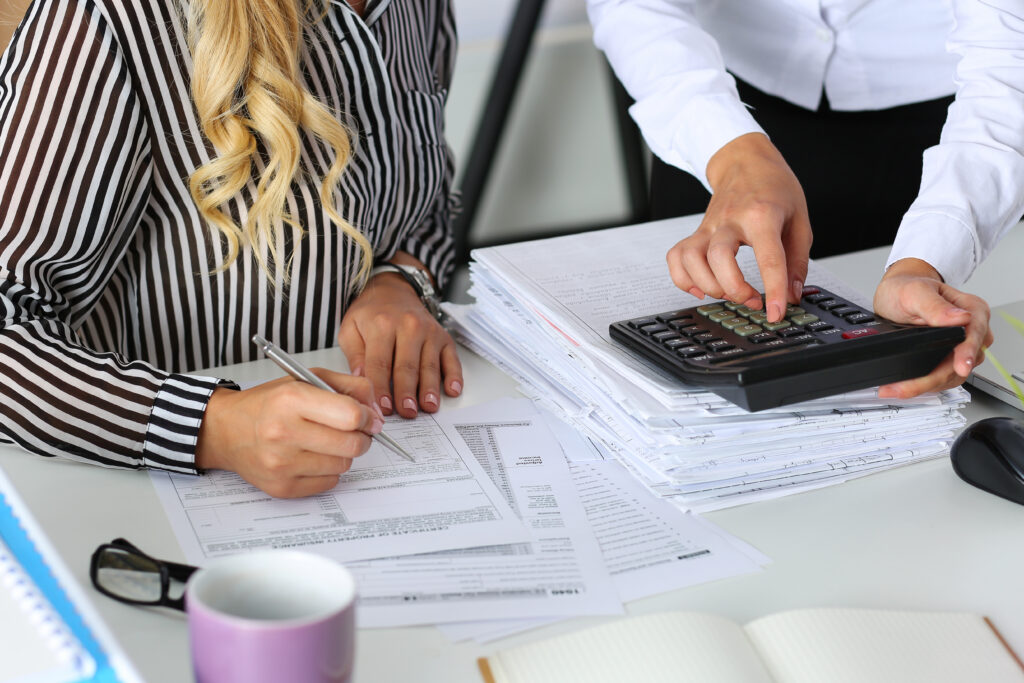 Two people working at a desk covered with documents and a large calculator. One person, wearing a striped shirt, writes on a nondeductible IRA form, while the other, in a white shirt, points at the calculator. A notebook and a mug are on the desk.