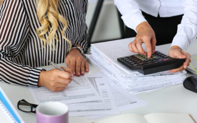 Two people working at a desk covered with documents and a large calculator. One person, wearing a striped shirt, writes on a nondeductible IRA form, while the other, in a white shirt, points at the calculator. A notebook and a mug are on the desk.