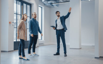 A real estate agent presents a spacious, empty apartment to a couple, highlighting its potential as a prime rental investment. The room boasts large windows, white walls, and a tiled floor. With a tablet in hand, the agent gestures upward to emphasize the high ceilings.