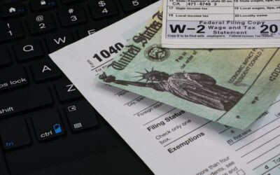 A federal income tax form and a W-2 wage statement rest on a black keyboard. Nearby, a United States Treasury check is partially visible, hinting at taxes for household employees among other tax-related financial documents.