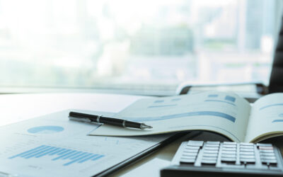 An open business report featuring charts and graphs sits on a desk, accompanied by a pen and calculator. In the background, city buildings blur through the window, perhaps reflecting on changes influenced by the Affordable Care Act.