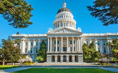 A grand white building with a large dome and columns stands proudly against a clear blue sky, surrounded by green trees and a well-kept lawn. Echoing the spirit of classic architecture, it symbolizes progress as California passes expansion in economic relief measures.