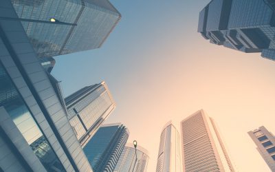 Looking up at a group of modern skyscrapers from street level, silhouetted against a clear sky with a warm, orange tint from the setting or rising sun, these glass and steel giants reflect the light. It's a dramatic urban scene emblematic of real estate market changes in 2022.