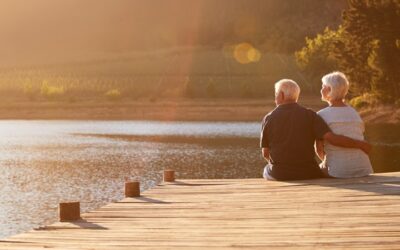 An elderly couple sits on a wooden dock embracing, gazing at a tranquil lake during sunset. Bathed in warm sunlight, they reflect on their journey together and the peace of retirement, with trees visible in the background—a testament to their dedication to the CalSavers Program.