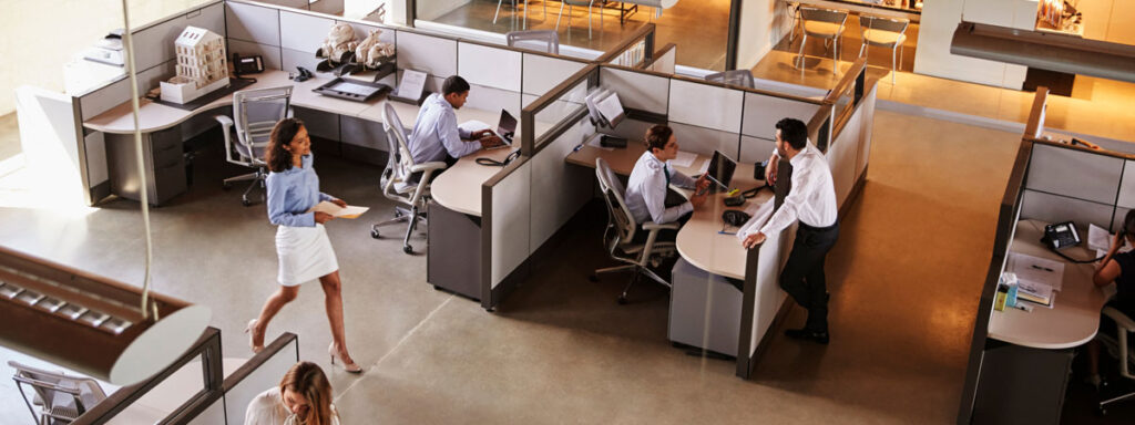 A modern office with cubicles buzzing with activity, where strategic use of space is evident. Two individuals focus on their computers, while another chats with a colleague. A woman in a blue shirt and white skirt navigates the well-lit area, embodying both energy and efficiency.
