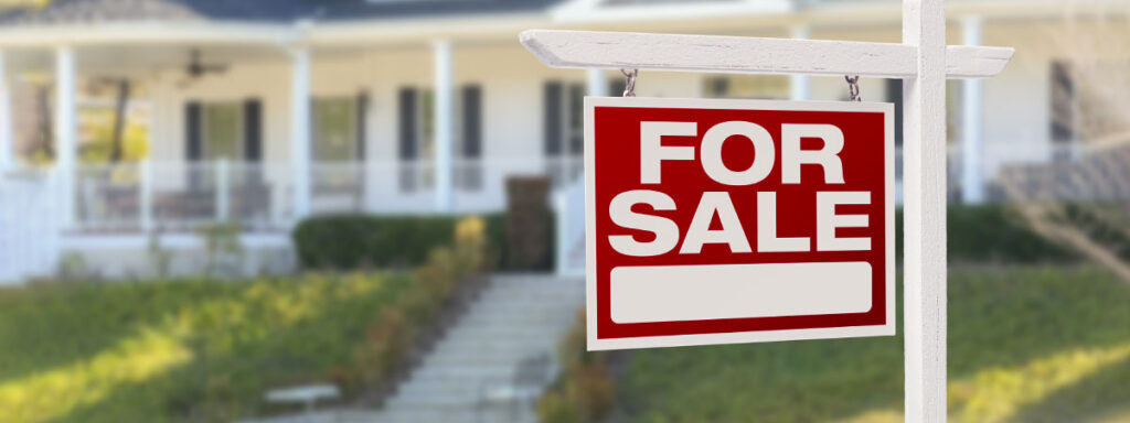 A "For Sale" sign with home sale details hangs on a white post in the foreground, set against a blurred view of a charming house. The porch, adorned with white railings and steps, blends seamlessly into the lush greenery surrounding it.