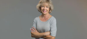 Head shot of middle-aged block woman wearing a casual grey blouse with her arms crossed. She is smiling.