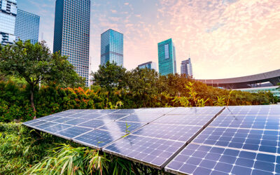Solar panels are installed in an urban green space with tall, modern skyscrapers and a pink-orange sky at sunset in the background. The scene captures the integration of renewable energy within a city environment.