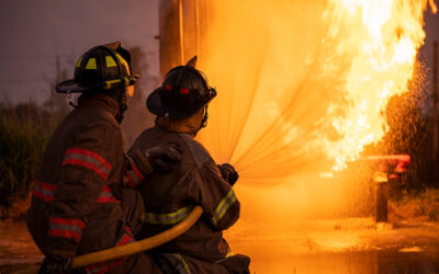 Two firefighters in protective gear kneel while aiming a hose at a large, intense fire. The scene is dramatic, with bright flames and smoke in the background—an all-too-familiar sight during a Los Angeles County disaster. The firefighters work diligently facing the blaze.